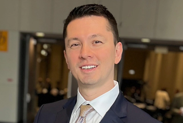 Professional headshot of smiling man in navy blazer and tie in dental office setting.