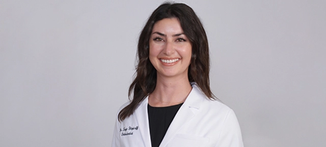 Professional headshot of a female dentist wearing a white coat and black shirt against a neutral background.