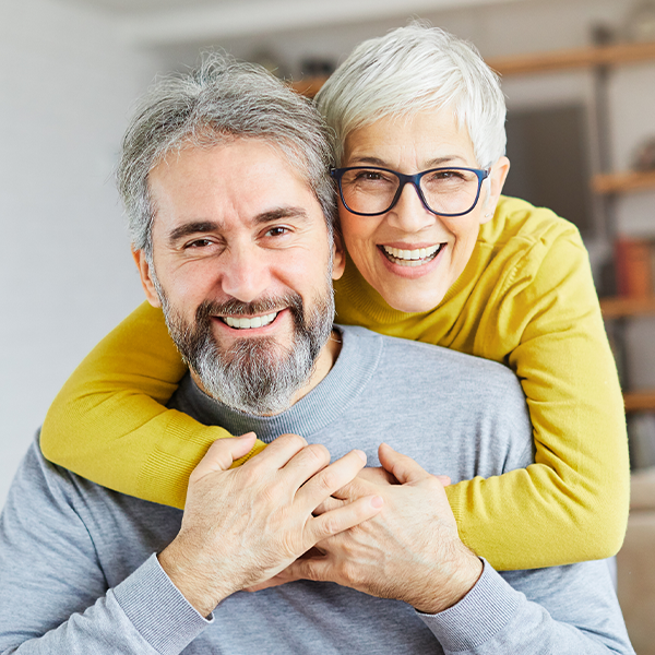 Happy mature couple smiling together indoors, man with beard wearing blue shirt, woman in yellow cardigan.