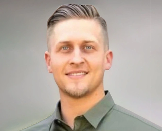 Professional headshot of smiling male staff member wearing olive green shirt against neutral background.