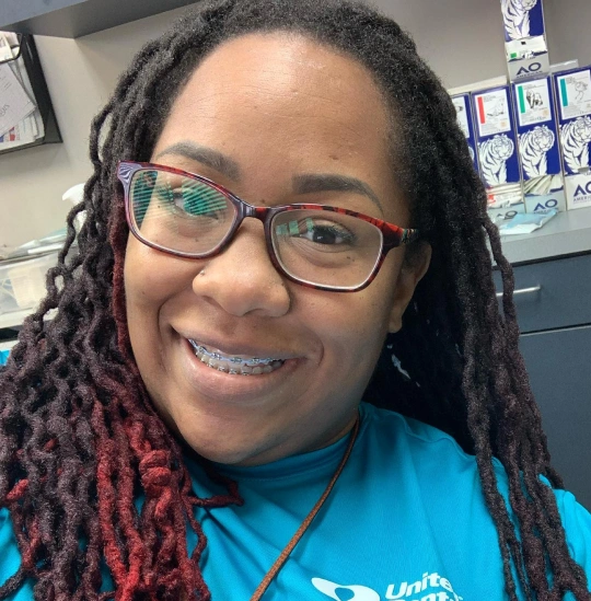 Smiling woman with braces and glasses wearing blue uniform in dental office setting.