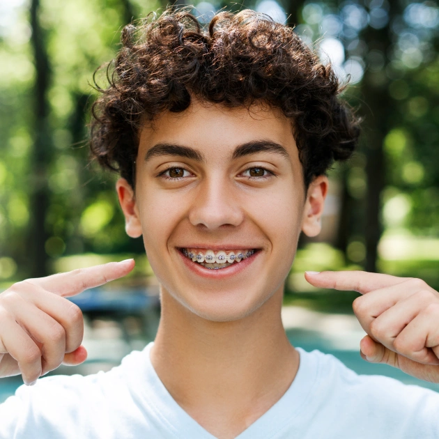 Smiling teenage boy with braces pointing to teeth outdoors in park setting