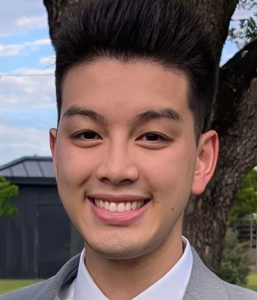 A close-up portrait of a smiling young Asian man outdoors. He has short, styled, upward-spiky black hair, neat eyebrows, and a slight beauty mark on his left cheek. He is wearing a white collared shirt under a gray suit jacket. The background is softly blurred, showing a large tree trunk and a dark building under a partly cloudy sky.
